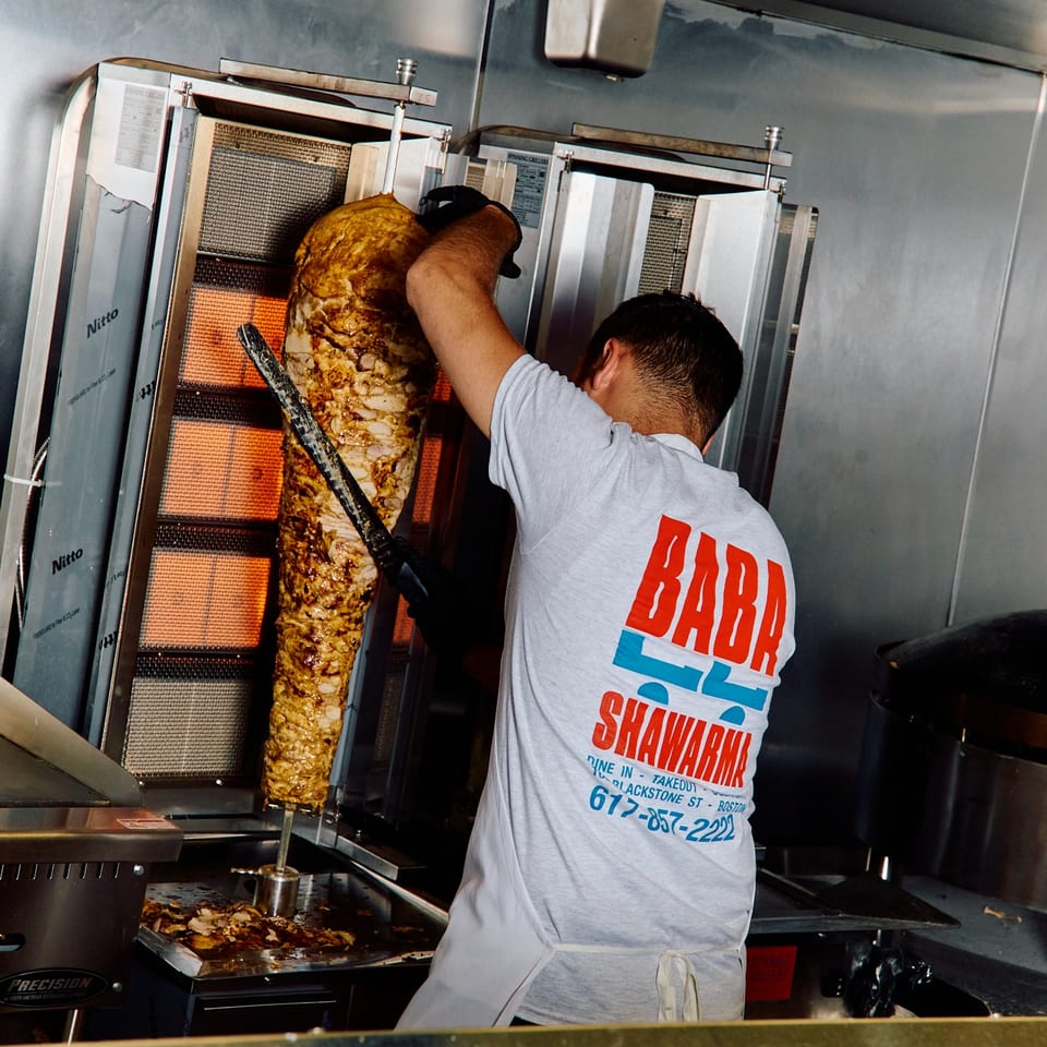 Chef in a "Baba Shawarma" shirt carving meat from a large vertical rotisserie.
