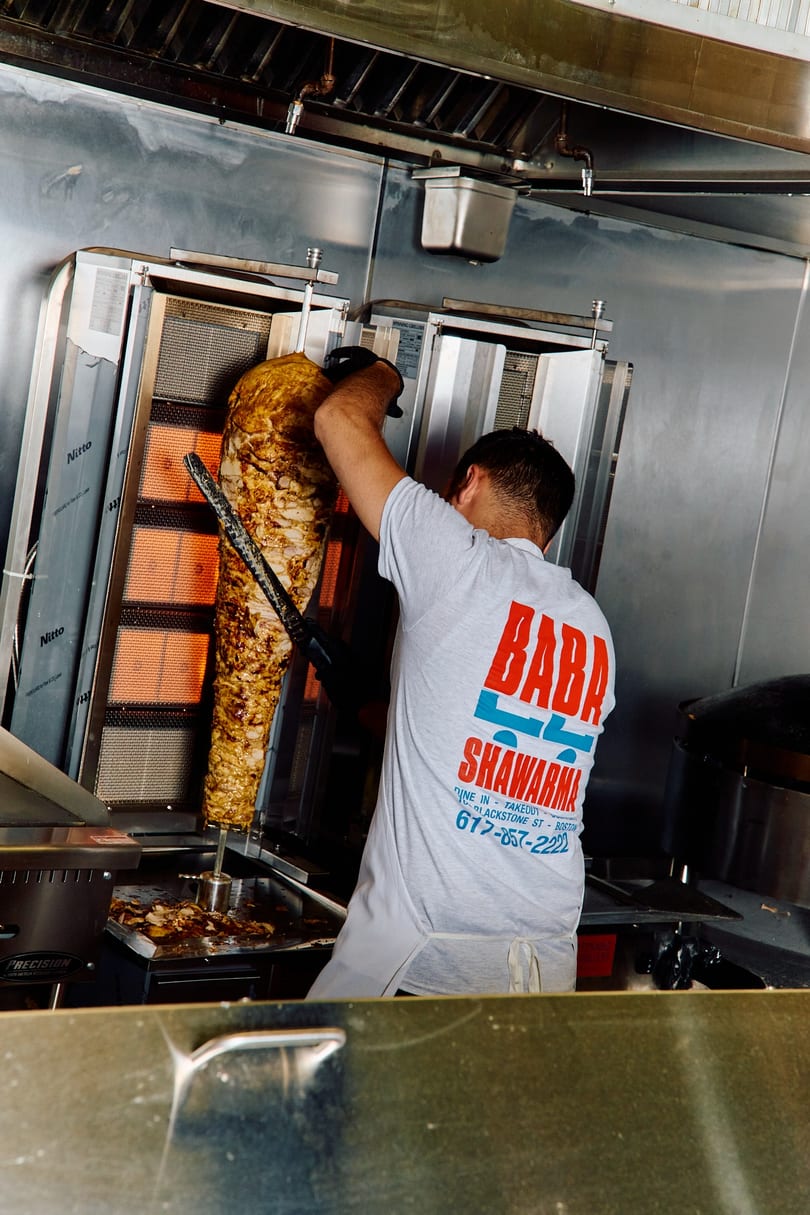 Chef in a "Baba Shawarma" shirt carving meat from a large vertical rotisserie.
