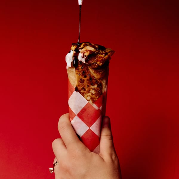 A hand holds a dessert crepe wrapped in red and white checkered paper, drizzled with chocolate sauce.